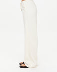 A person in loose, light-colored BOW N ARROW NOVA FLARE PANT by The Upside and black sandals stands in profile against a plain white background.