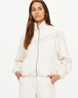 A woman with long brown hair wears The Upside’s BOW N ARROW FRINGE ZIP JACKET, a light-toned jacket with geometric design and fringe, paired with matching pants, standing hands-in-pockets against a plain white background.