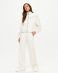 A woman with long brown hair stands confidently against a white background, wearing The Upside's BOW N ARROW FRINGE ZIP JACKET and matching wide-leg pants with white sneakers. Her hands are in her pockets as she looks at the camera.