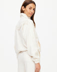 A woman with long brown hair wears The Upside's BOW N ARROW FRINGE ZIP JACKET, an off-white piece with a high collar and subtle geometric design, standing sideways and looking over her shoulder against a white background.