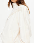 A woman is shown from the side against a plain background wearing The Upside's BOW N ARROW FRINGE ZIP JACKET in off-white, featuring subtle jacquard fabrication, textured patterns, and fringe detailing.