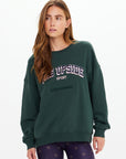 A woman with long brown hair wears The Upside’s ALUMNI COOLUM CREW organic cotton sweatshirt in dark green and patterned black leggings, standing against a plain white background.