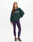 A woman with long, loose hair and a neutral expression stands against a white background wearing The Upside's ALUMNI COOLUM CREW sweatshirt in dark green, navy leggings with a small pattern, and brown sneakers with white soles.