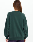 A person with long brown hair faces away, wearing The Upside's ALUMNI COOLUM CREW sweatshirt in dark green and patterned dark leggings, standing against a plain white background.