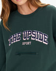 A person wears The Upside’s ALUMNI COOLUM CREW: a dark green organic cotton sweatshirt with “THE UPSIDE” in bold white and pink, “SPORT” below, classic vintage collegiate style, and a small horizontal zippered pocket under the text.