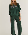 A woman sports The Upside's ALUMNI COOLUM CREW, a loose dark green organic cotton sweatshirt with "THE UPSIDE SPORT" print, paired with matching green wide-leg pants featuring white side stripes and gold bracelets, evoking vintage collegiate style.