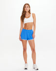 A woman with long brown hair and a neutral expression stands against a white background, wearing The Upside's WRANGLER CALLIE SHORT in azure blue, a white sports bra, and white sneakers.