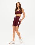A woman wears The Upside's TRAIL 6IN SPIN SHORT, a burgundy high-waisted short with white stripes and matching sports bra, paired with white athletic shoes. This breathable set is made from recycled materials for comfort and sustainability.