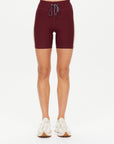 Someone stands against a plain white background wearing The Upside TRAIL 6IN SPIN SHORT—maroon athletic shorts with a white drawstring and side stripes, made from breathable, moisture-wicking fabric—paired with white sneakers.
