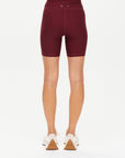 A person, visible from the waist down and facing away, wears The Upside's TRAIL 6IN SPIN SHORT in maroon—high-waisted, breathable athletic shorts—paired with white sneakers against a white background.