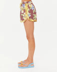 A person stands in profile wearing The Upside ISLA KLIO SHORT with a brown floral print and yellow trim, paired with light blue platform sandals with yellow thong straps, set against a plain white background.