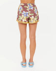 A person stands facing away, wearing The Upside ISLA KLIO SHORT in vintage hibiscus—brown with yellow trim—and blue sandals. The shorts are made from a soft lyocell-cotton blend. Background is plain white.