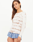 A woman with long brown hair wears The Upside AYLA CHRISSY LONG SLEEVE TOP, styled with blue-and-white striped shorts, standing against a plain white background.