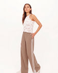 A woman with long brown hair poses confidently against a white backdrop, wearing The Upside’s JULIET PANT S26 with white side stripes, a sleeveless tie-dye tank top, and white sneakers. One hand rests on her hip.