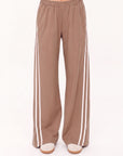 A person wears The Upside JULIET PANT S26—loose, light brown twill pants with two vertical white side stripes, elasticated waist, and wide legs—standing against a plain white background.