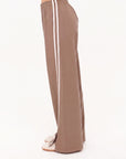 A person stands sideways against a plain white background, showcasing only their lower body and feet in The Upside JULIET PANT S26—loose, lightweight twill pants with an elastic waist and two white side stripes.
