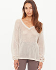 A woman with long brown hair wears The Upside's JOSEPHINE V NECK KNIT—a cream, open-knit relaxed sweater—paired with patterned leggings, standing against a white background and gazing neutrally at the camera.