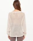 A woman with long brown hair faces away, wearing The Upside's JOSEPHINE V NECK KNIT—a loose, cream open-knit sweater with a relaxed V neckline—paired with patterned brown leggings against a plain white background.
