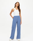 A woman stands against a plain white background wearing The Upside’s SIERRA PANT in light blue, paired with a white sleeveless top and white shoes, one hand in her pocket and a neutral expression.