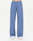 A person wears The Upside SIERRA PANT—loose-fitting, light blue, moisture-wicking wide-leg pants with an elastic waistband—and white shoes, shown waist down on a plain white background; ideal for comfortable travel days.