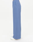 A person stands in profile wearing The Upside's SIERRA PANT, loose-fitting wide leg pants made from recycled polyamide with an elastic waistband and side pockets, paired with white shoes against a plain white background.