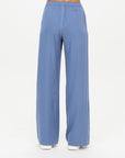 A person wears The Upside's SIERRA PANT in light blue recycled polyamide, featuring a wide leg and elastic waistband. Moisture wicking fabric ensures comfort, paired with white shoes against a plain background that emphasizes the relaxed fit.