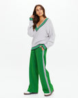 A woman stands against a white background wearing The Upside's wide-leg GALAXIA LORETTA PANT in green with white and navy side stripes, paired with a loose gray cable-knit sweater featuring Amazon Green trim. She looks to the side, hand near her chin.