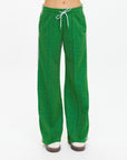 A person wearing The Upside GALAXIA LORETTA PANT in Amazon Green, featuring a drawstring waist and white side stripes, stands against a white background with only the lower torso and legs visible.