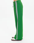 Person wearing The Upside GALAXIA LORETTA PANT in Amazon Green with white and black side stripe, paired with white socks and brown shoes, standing sideways against a plain white background.
