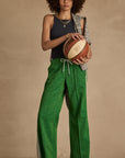 Wearing a black tank top and The Upside's GALAXIA LORETTA PANT in Amazon Green, a person with curly hair stands against a neutral background, holding a basketball with a gray sweater draped over their shoulder.