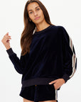 A woman wears The Upside MONACO LISEL CREW NECK SWEATER in navy velour with striped sleeves and matching recycled polyester shorts, standing against a white background and looking at the camera with a relaxed expression.