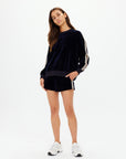 A woman stands against a white background wearing The Upside’s MONACO HELENA SHORT in navy velour with matching striped sweatshirt, creating a relaxed, casual look paired with her long brown hair and white sneakers.