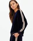 A woman with long brown hair stands sideways in The Upside MONACO LISEL CREW NECK SWEATER, a navy velour sweatshirt made from recycled polyester with beige and white striped sleeves, paired with matching shorts. She looks slightly toward the camera.