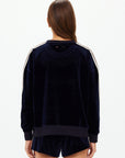 A woman with long brown hair, seen from behind, is wearing The Upside’s MONACO LISEL CREW NECK SWEATER in navy velour with white sleeve stripes and matching recycled polyester shorts, set against a plain white background.