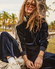 A woman with long hair sits outdoors near palm trees, wearing The Upside's MONACO LISEL CREW NECK SWEATER, sunglasses, white sneakers, and gold jewelry. Headphones rest around her neck as she poses in a relaxed, casual style.