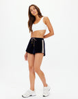 A woman with long brown hair poses confidently in The Upside's MONACO HELENA SHORT—relaxed fit black athletic shorts with side stripes made from recycled polyester—paired with a white sports bra and sneakers, against a white background.