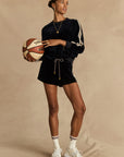 A woman in The Upside's MONACO HELENA SHORT, navy velour athletic outfit, white sneakers, and gold jewelry holds a basketball against a beige backdrop, looking to the side.