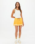 A woman stands facing forward wearing The Upside’s PRIX QUINNY PLEATED SKIRT—a mimosa yellow mini with striped drawstring—paired with a white sleeveless top, white socks with yellow stripes, and white sneakers against a plain white background.