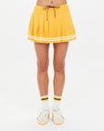 A person wears The Upside's PRIX QUINNY PLEATED SKIRT in mimosa yellow with white stripes, paired with matching yellow and white sneakers and white socks featuring yellow and black stripes, standing against a plain white background.