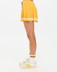 A person stands sideways in The Upside PRIX QUINNY PLEATED SKIRT, mimosa yellow with white stripes, paired with white socks with yellow and black stripes and white Adidas sneakers with yellow stripes, set against a plain white background.