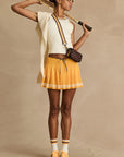 A woman in a sporty outfit poses with a tennis racket, wearing The Upside's PRIX QUINNY PLEATED SKIRT in mimosa yellow, a white sleeveless top, yellow sneakers, striped socks, sunglasses, a crossbody bag, and a sweater over her shoulders.