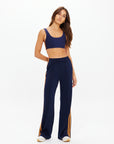 A woman in The Upside VITESSE MARNIE WIDE LEG PANT, featuring a high rise and elasticated waistband, stands against a white background with one hand behind her back and her head slightly tilted.