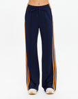 A person wears The Upside VITESSE MARNIE WIDE LEG PANT—navy with orange side stripes, high rise and wide leg fit, drawstring waist—with white sneakers, standing against a plain white background.