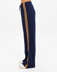 A person in The Upside VITESSE MARNIE WIDE LEG PANT—high rise, wide leg, navy with orange side stripes and elastic waistband—paired with white sneakers, shown in profile against a plain white background.