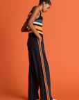 A woman stands sideways against an orange background, wearing The Upside VITESSE MARNIE WIDE LEG PANT with orange side stripes and a sleeveless striped knit top. She pairs the look with white sneakers, hands in her pockets, hair in a bun.