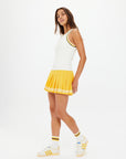 A woman stands sideways against a white background, wearing The Upside's KIRSTY RIB TANK TOP, a yellow pleated skirt with white and yellow stripes, plus white sneakers and matching socks. She looks at the camera.
