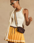 A woman wearing The Upside’s KIRSTY RIB TANK TOP, white sunglasses, a yellow pleated skirt, and a checkered crossbody bag with a sweater over her shoulder stands against a beige background.