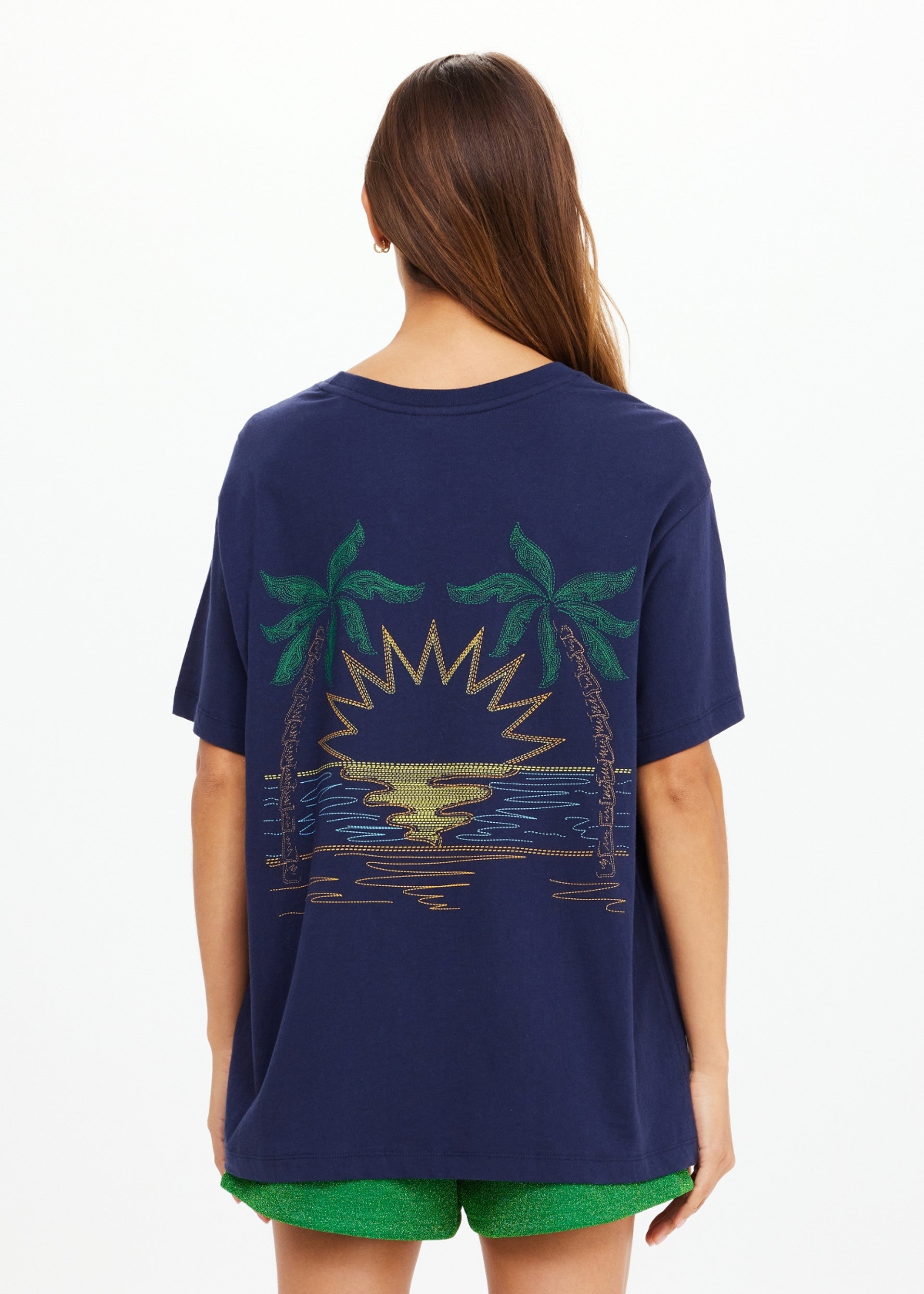 A woman with long brown hair, seen from behind, wears The Upside SAMMI TEE—a navy graphic t-shirt with palm trees and a sun over water—paired with bright green shorts.