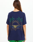 A woman with long brown hair, seen from behind, wears The Upside SAMMI TEE—a navy graphic t-shirt with palm trees and a sun over water—paired with bright green shorts.