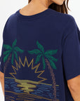 A woman wears The Upside’s SAMMI TEE, an oversized navy graphic tee with an embroidered palm tree, sunset, and water design on the back. Only her back and part of her face are visible.